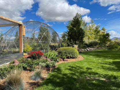 An idyllic backyard scene from a home in Washington state hosts a large 28 Ft aviary.  The dome is surrounded by an appealing selection of plantings and bordered by a lawn of deep green.